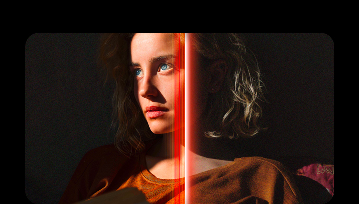 A woman with piercing blue eyes and a burnt orange top in a dark space. Red lines depicting AI refinements cover part of her face, which is bright and detailed, while the rest of the image looks less colorful.	
