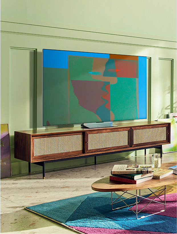 A swivel stand LG OLED television is in a colorful living room with wooden furnishing and greenery. A swivel stand LG OLED television is in a gray and abstract room.