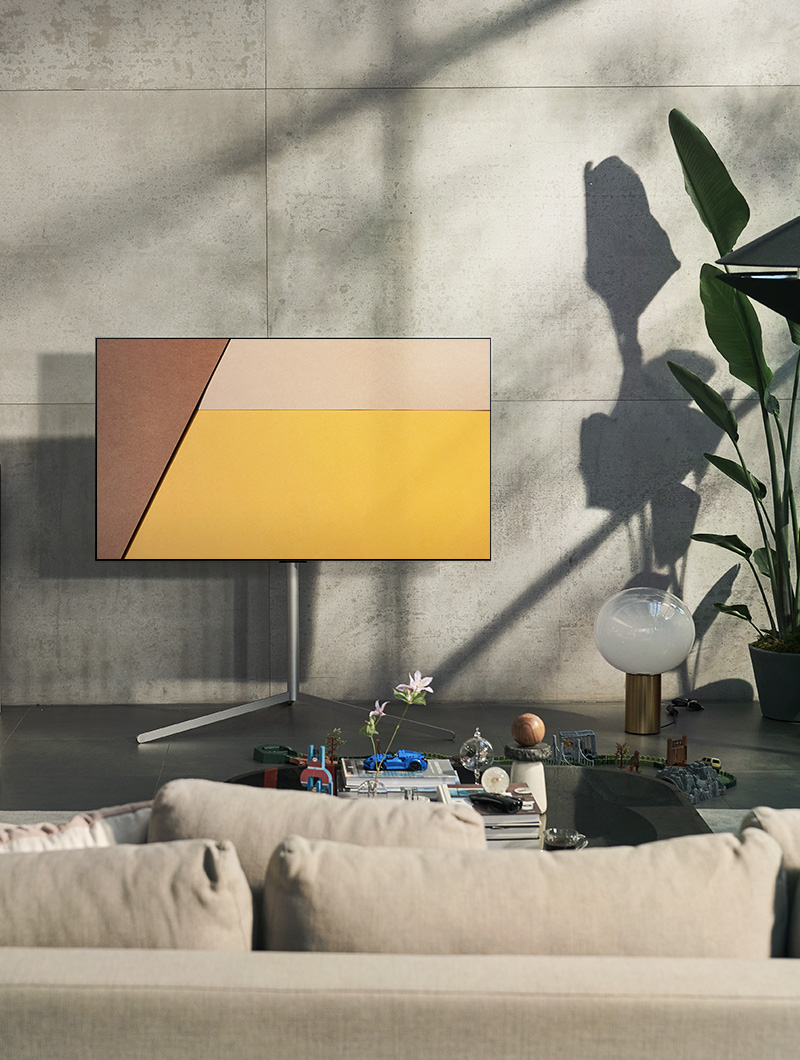 An LG OLED television is in a modern gray living space against a window showing a swimming pool. An LG OLED television is in a neutral living space with exposed bricks, wooden details, and greenery.