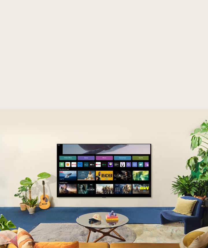 A whale floating above the ocean with a woman in the ocean are shown. A home screen appears from below. As the scene transitions, it shows the image in a large LG TV on the wall. Two women sit in a cozy and neutral living room filled with plants, and a guitar. One woman points the remote at the TV, which shows a range of apps and recommended TV shows on the home screen. 