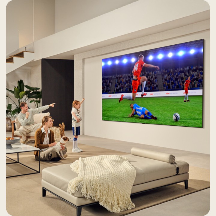 A family is in their living room. They have a wall-mounted LG TV with an ultra big screen that's almost as huge as the whole wall. They're watching an exciting soccer game on the large and immersive screen.  
