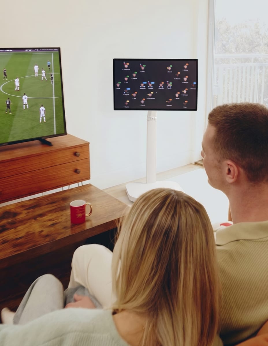 The back of a man and a woman sitting side by side on a sofa, watching a football match on the TV while checking a football match analysis screen on an LG Smart Monitor Swing.
