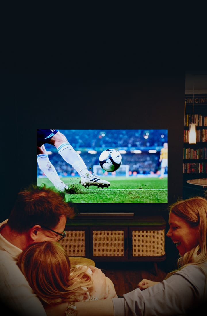 A family in a cozy living room watches a soccer match on a large wall-mounted TV, showing a close-up of a player kicking a ball on a stadium field, with warm lighting and a cinematic atmosphere.