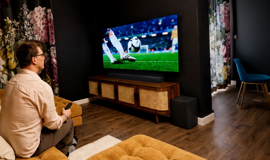 A man sits on a sofa in a bright living room, holding a game controller while watching a soccer match on a wall-mounted TV.