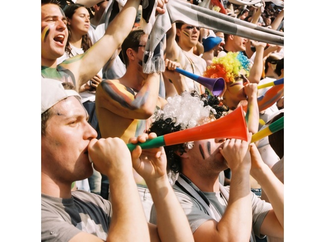 A group of enthusiastic fans in a stadium cheer and blow colorful vuvuzelas, wearing team colors and raising their arms in excitement.