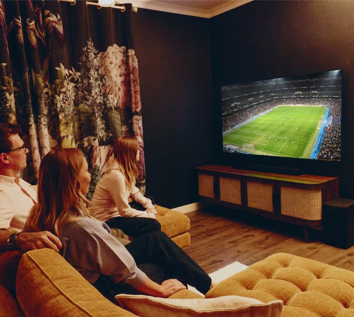 A family of three sits together on a sofa in a cozy, dimly lit living room, watching a soccer match on a large LG OLED TV. The room features dark walls, floral curtains, a wooden media console, and speakers, creating a warm and intimate atmosphere.