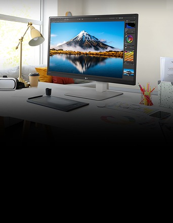 On the desk, there is an UltraFine monitor displaying Photoshop work. Next to the monitor, there is a desk lamp, a mouse, paper, and coloured pencils.