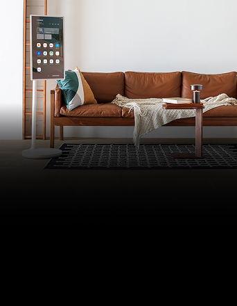LG StanbyME, a vertical smart display, stands next to a leather sofa in a modern living room, showing app icons and widgets on screen.