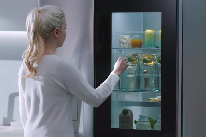 Woman with blonde hair uses LG fridge with glass panel, revealing beverages and snacks inside.