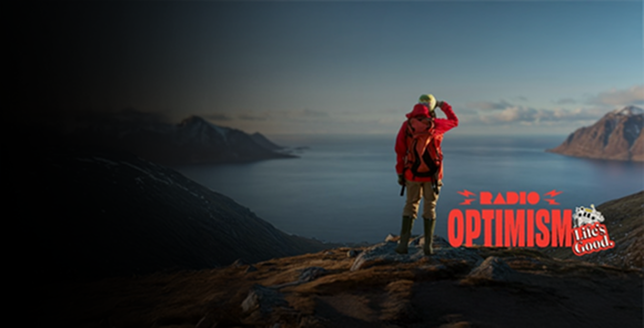 A woman wearing a red hiking jacket is standing in a place where the vast ocean and snow-covered mountains can be seen. The image also features the "Radio Optimism" slogan logo and the "Life's Good." logo alongside the woman.
