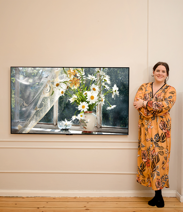 The video sequentially shows the woman standing next to a TV with a vase placed by a sunlit window and smiling and the woman standing next to a washing machine.