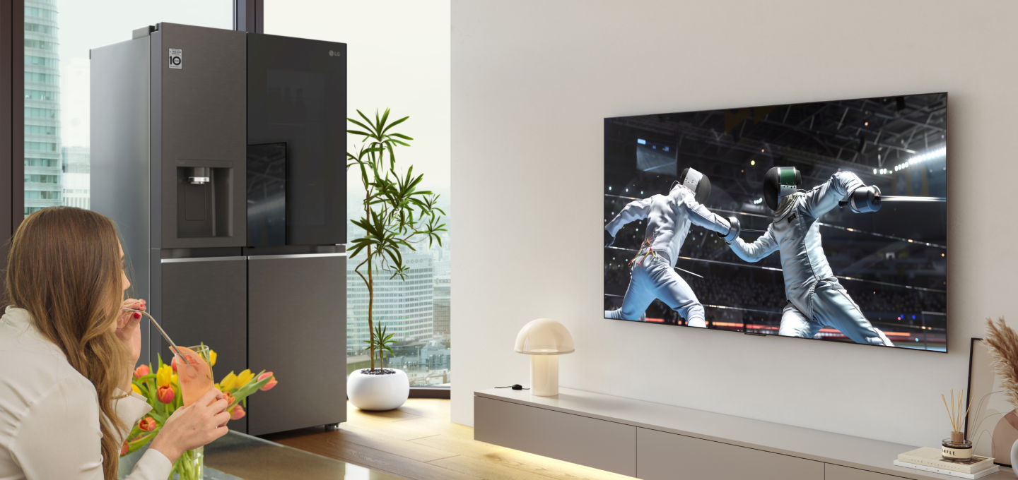 A woman watching a sports game on an LG OLED TV in the living room, holding a mocktail, with an LG InstaView refrigerator next to the TV wall.