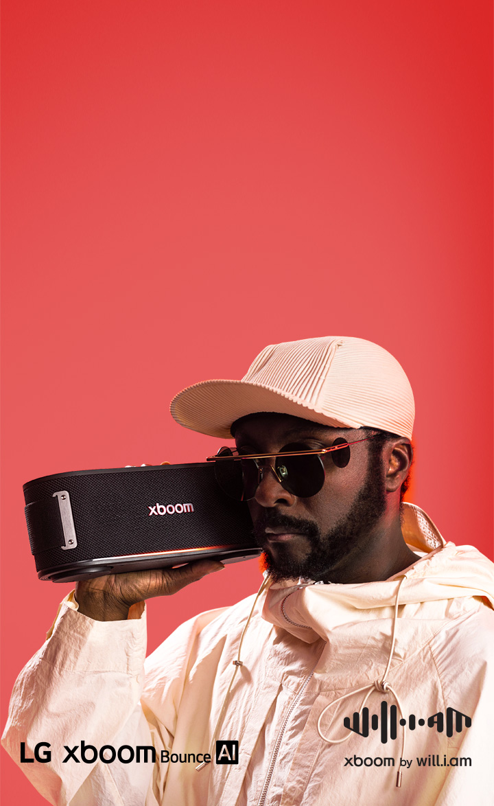will.i.am holds an xboom speaker to his ear, with an orange sun set colour background