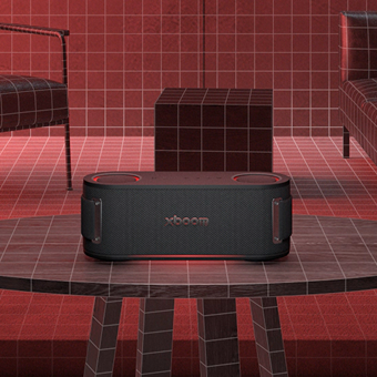 LG XBOOM speaker placed on a table in a red-toned room with grid-patterned walls and modern furniture