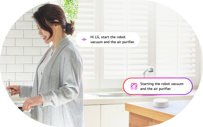 An image of the LG Smart Speaker AI voice command feature. A woman works in the kitchen while voice commands and responses are shown in speech bubbles. Command: 'Hi LG, turn on the robot vacuum and air purifier.' Response: 'Turning on the robot vacuum and air purifier.'