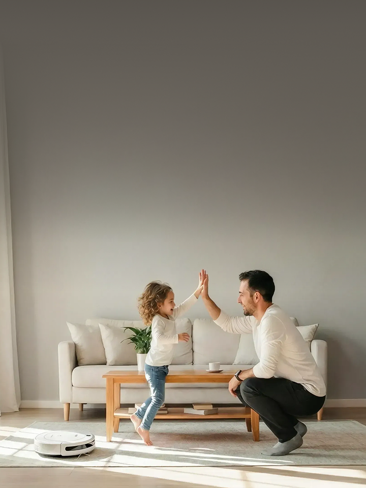 A smiling man and a child standing on a coffee table high-fiving in a bright living room, with a robot vacuum on the floor